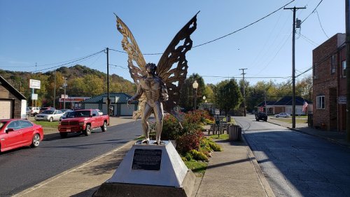 A large metal butterfly sculpture stands on a sidewalk, surrounded by flowers and a small town street in the background.