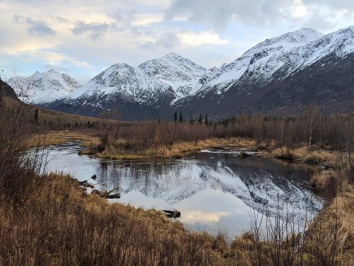A serene landscape featuring a river reflecting snow-capped mountains and autumn foliage under a cloudy sky.