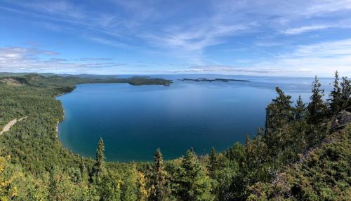 A panoramic view of a serene blue lake surrounded by lush green forests and distant islands under a clear sky.