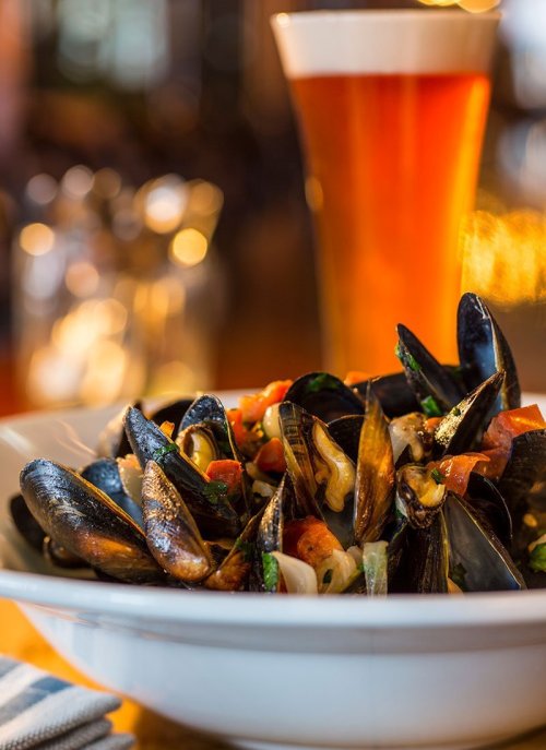 A bowl of mussels with tomatoes and herbs, accompanied by a tall glass of amber beer in the background.