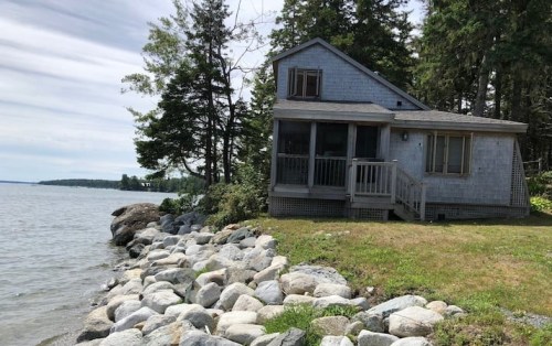 A cozy house by the water, surrounded by trees and rocky shoreline under a cloudy sky.