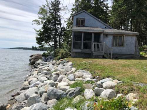 A cozy house by the water, surrounded by trees and large rocks along the shoreline on a sunny day.