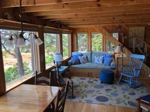 Cozy cabin interior with a blue couch, rocking chair, and large windows overlooking nature. Light wood accents throughout.