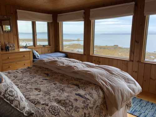 Cozy bedroom with a bed, wooden walls, and large windows overlooking a calm lake and distant shoreline.