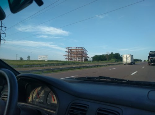 A view from a car on a highway, showing a partially constructed building and green fields under a blue sky.