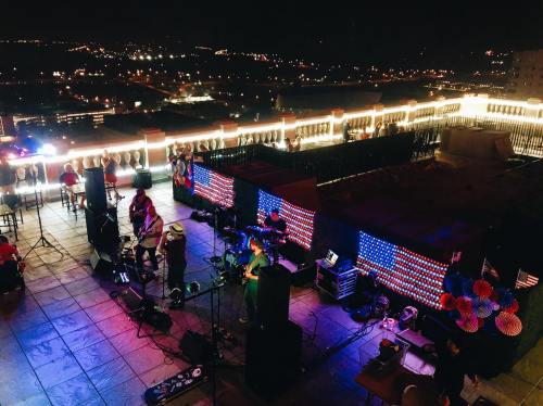 A rooftop concert at night with a band performing, colorful lights, and a city skyline in the background.