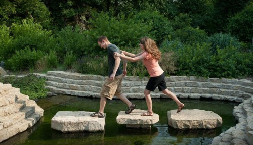 A man and woman playfully cross stepping stones over a small pond in a lush, green garden.