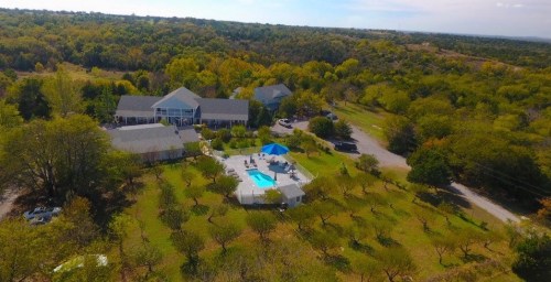 Aerial view of a property with a pool, surrounded by trees and open land, featuring several buildings in the background.
