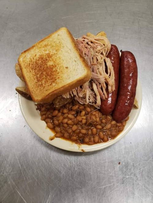 A plate with pulled pork, two sausages, baked beans, and a slice of toasted bread.