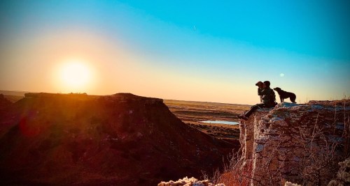 A person and a dog sit on a cliff, watching a vibrant sunset over a vast landscape.