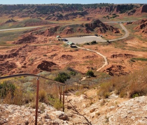 A view of a red, rocky landscape with a winding path and a small area of buildings in the distance.