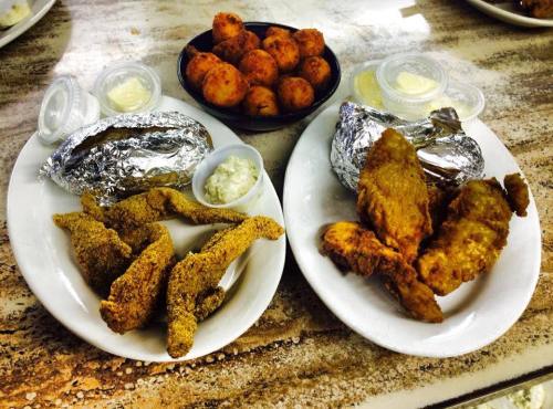 Two plates of fried fish and chicken, with sides of baked potatoes and hushpuppies, served with dipping sauces.