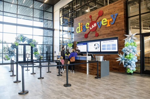 Interior of a modern discovery lab with a colorful sign, reception area, and balloon decorations. Visitors are checking in.
