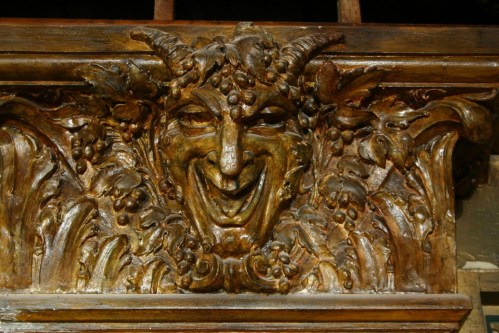 Carved wooden relief of a smiling, horned figure surrounded by foliage and grapes, showcasing intricate detailing.