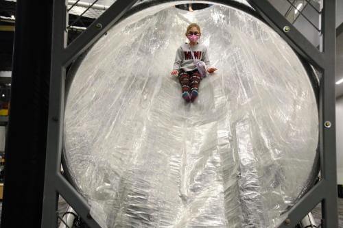 A child sits inside a large, transparent, plastic-covered structure, wearing a mask and colorful clothing.
