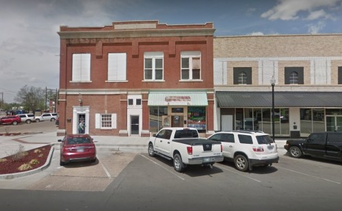 A red brick building with white shutters, surrounded by parked cars in a small town setting.