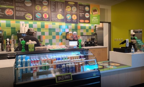 A colorful cafe interior with a display case of drinks, a menu board, and staff preparing food in the background.