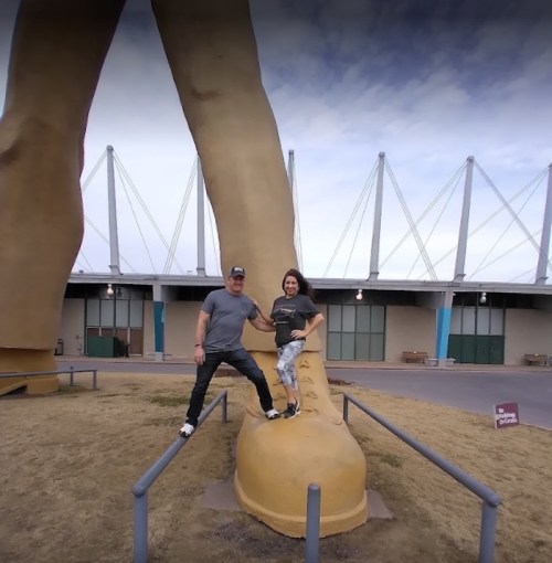 A man and woman pose on a giant shoe sculpture, with large legs in the background and a building behind them.