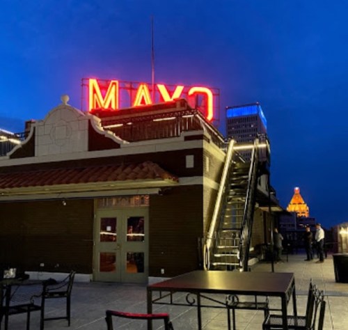 Rooftop view at night featuring a bright "MAYO" sign and city skyline in the background.