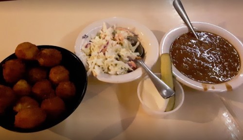 A bowl of golden fried balls, a side of coleslaw, and a dish of brown beans with pickles on the side.