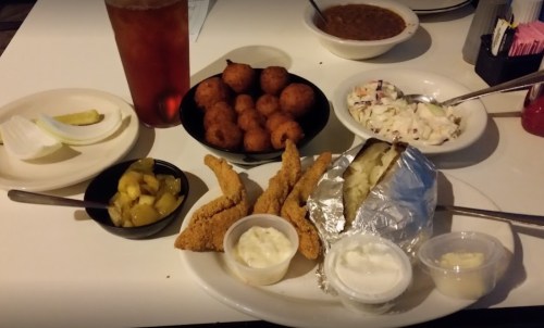 A plate of fried fish, coleslaw, pickles, baked potato, hushpuppies, and a drink, with a bowl of chili in the background.