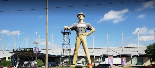 A large statue of a man in a hard hat stands outside an expo center, with blue skies and clouds in the background.