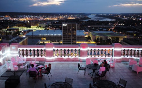 Rooftop view of a city at dusk, featuring outdoor seating and illuminated buildings in the background.