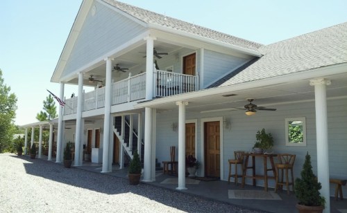 A light blue two-story building with a porch, columns, and outdoor seating, surrounded by gravel and greenery.