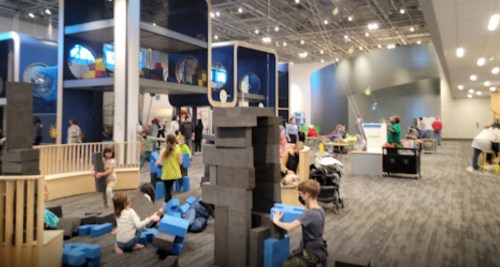 Children play with large building blocks in a spacious, colorful activity area filled with families and interactive exhibits.