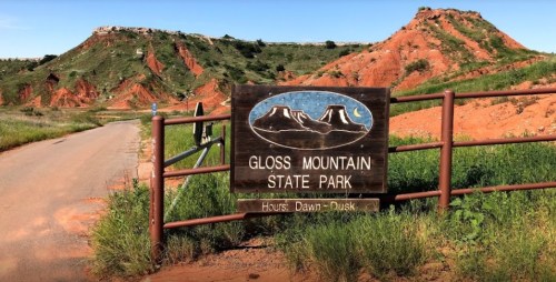 Sign for Gloss Mountain State Park with red rock formations and green grass in the background, under a clear blue sky.