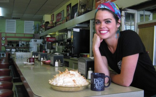 A smiling woman with a colorful headband sits at a diner counter next to a large meringue pie and a coffee mug.