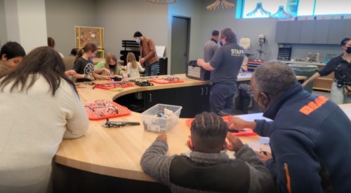A group of people, including children and adults, work together at a table with tools and materials in a workshop setting.