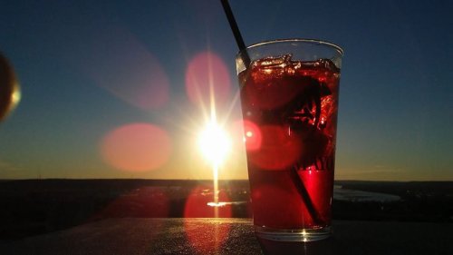 A glass of red drink with ice and a straw, silhouetted against a vibrant sunset.