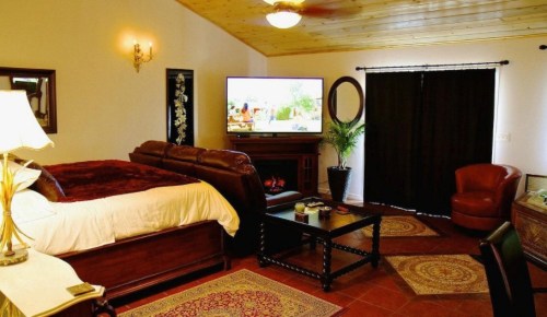 Cozy bedroom with a large bed, TV, armchair, and decorative rugs, featuring warm lighting and dark curtains.