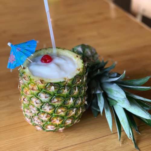 A fresh pineapple cup filled with a tropical drink, topped with a cherry and a small umbrella, on a wooden surface.