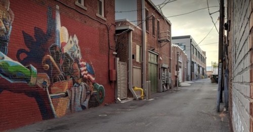 A narrow alleyway with a colorful mural on one wall and buildings lining both sides under a cloudy sky.