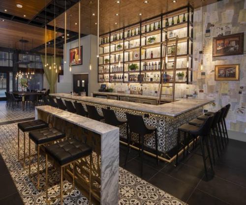 A stylish bar with a marble counter, black stools, and shelves lined with bottles, set in a modern restaurant interior.