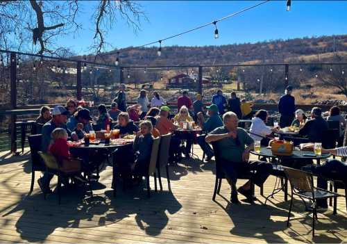 A lively outdoor dining area with people enjoying meals and drinks, surrounded by trees and hills on a sunny day.