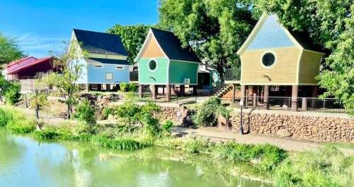 Colorful houses with unique designs by a serene waterway, surrounded by lush greenery and clear blue skies.