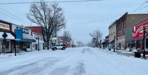 A snowy, empty street lined with shops and trees, with a light dusting of snow covering the ground and buildings.