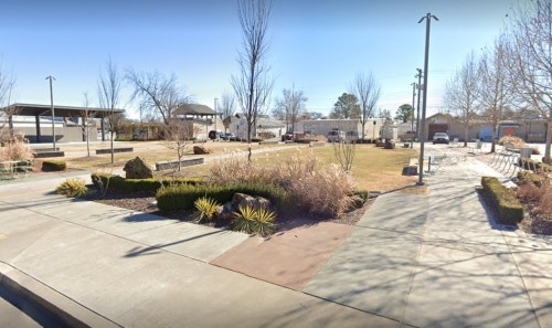 A spacious park with grassy areas, trees, and pathways, featuring benches and a few parked cars in the background.