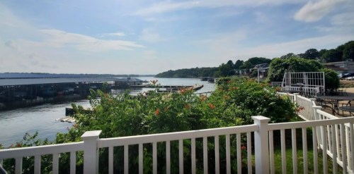 A serene waterfront view with boats, greenery, and a clear blue sky, framed by a white fence.