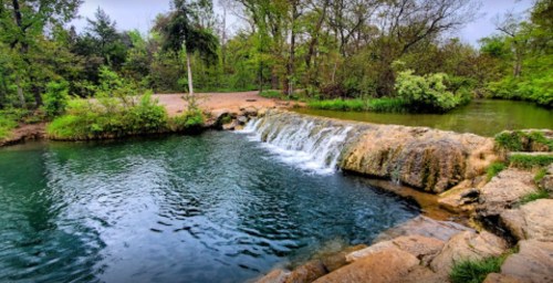 A serene waterfall cascades into a clear, greenish-blue pool, surrounded by lush trees and greenery.