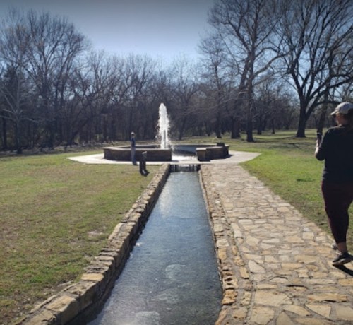 A sunny park scene featuring a fountain, a stone path, and trees in the background. Two people are walking nearby.