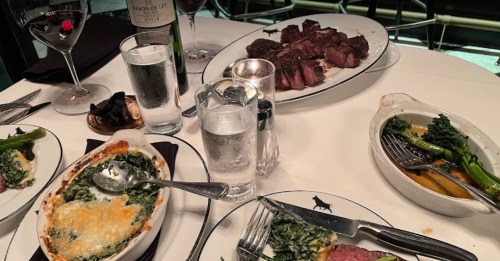 A dining table with plates of steak, creamed spinach, and asparagus, accompanied by glasses of wine and water.