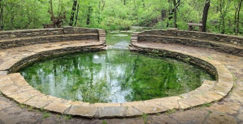 A circular stone pool surrounded by lush greenery and trees, reflecting the calm water and serene nature.
