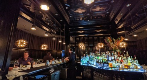 A stylish bar with a well-stocked shelf of colorful bottles, dim lighting, and patrons enjoying drinks.