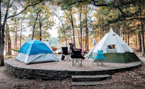 A campsite with two tents, chairs, and a hammock surrounded by trees and a stone circle.