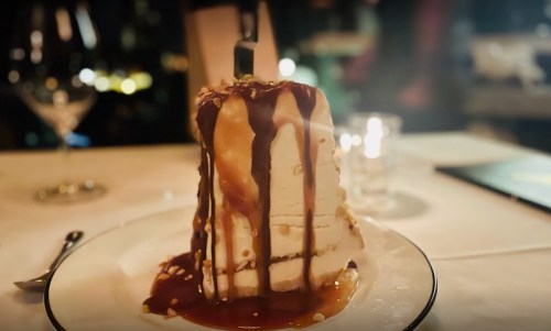 A tall slice of cake drizzled with caramel sauce, served on a white plate with a fork beside it.
