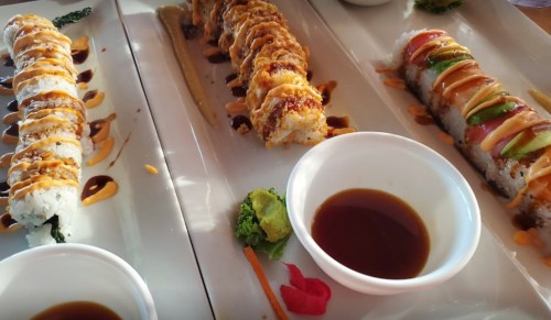 Three sushi rolls on a white platter, drizzled with sauces, accompanied by small bowls of soy sauce and wasabi.
